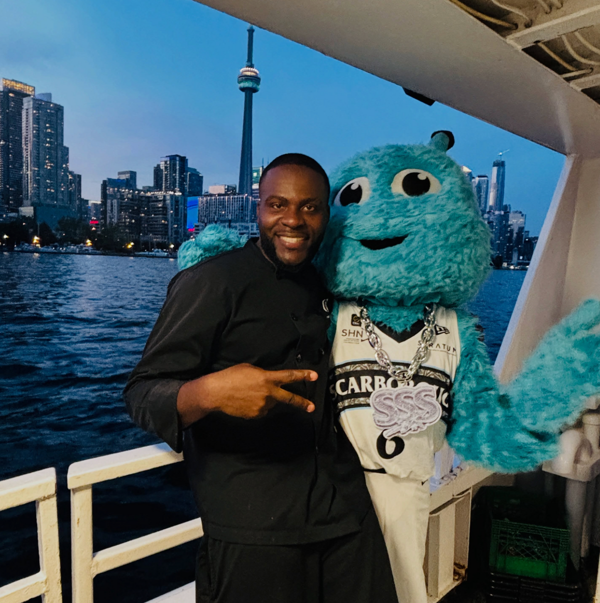 A man in a black shirt posing with a large blue monster mascot on a boat, with the Toronto skyline and CN Tower visible in the background at dusk.